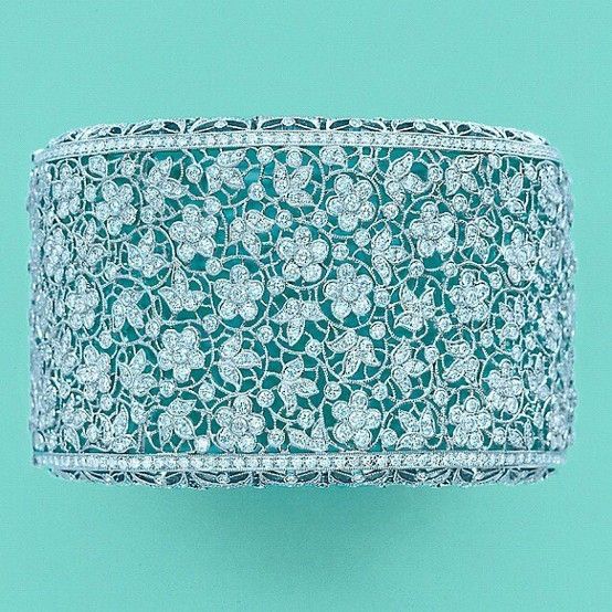 this is a closeup image of a silver diamond bracelet. the bracelet is adorned with a delicate floral pattern and is set against a backdrop of a bright turquoise surface. the sparkle of the diamonds and intricate craftsmanship of the bracelet are highlighted in this image.