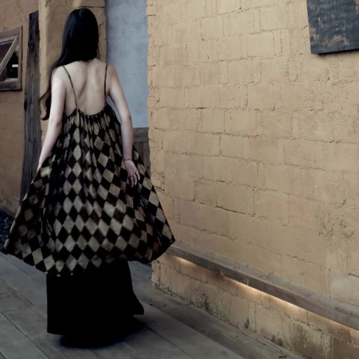 a woman is walking away from the viewer down a walkway indoors. she is wearing a dress with a black top and a diamondpatterned bottom, held up by spaghetti straps. the background consists of a brick wall.