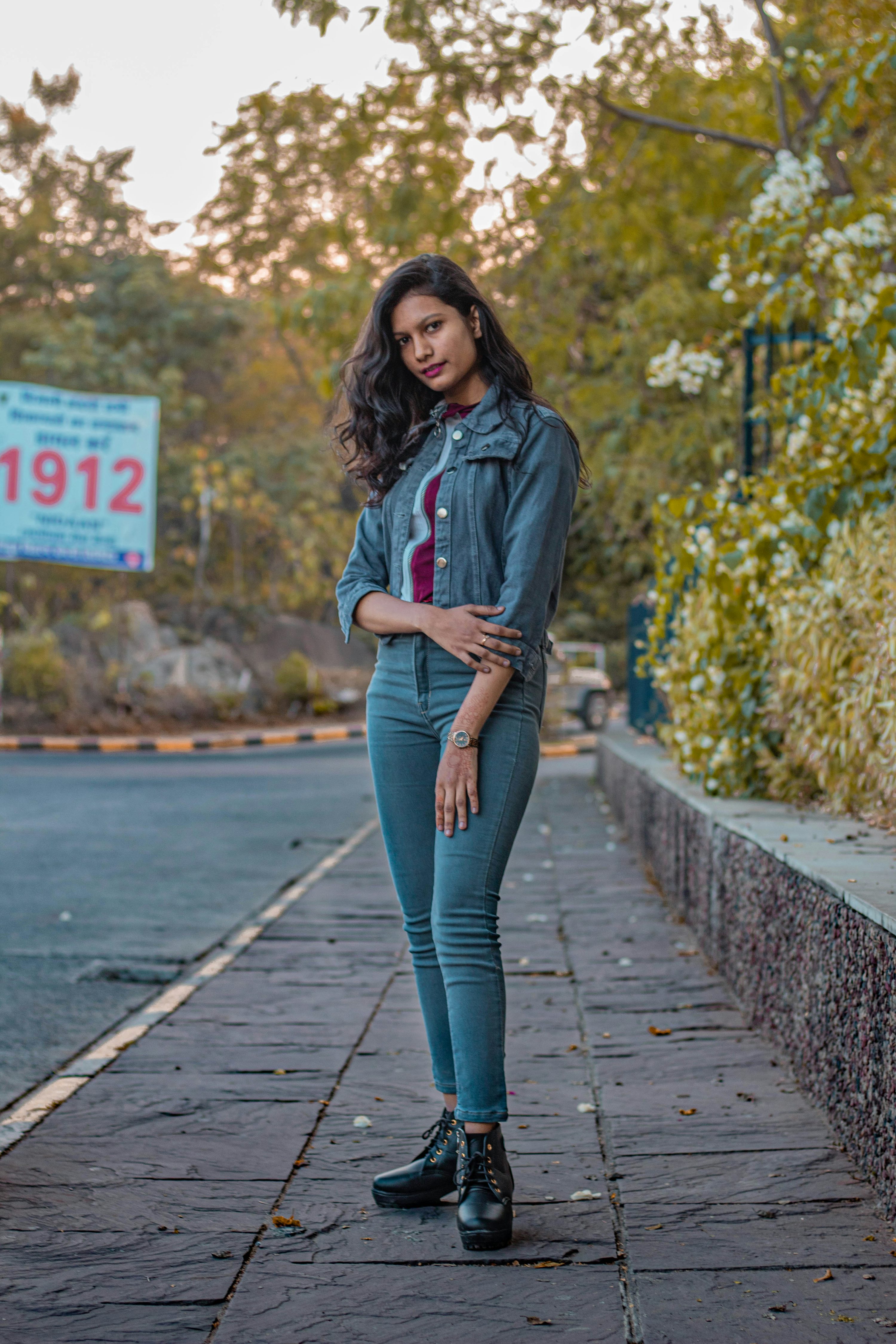 a fullbody shot of a young woman posing confidently on a city sidewalk.  she's wearing a teal denim jacket over a burgundy longsleeved shirt, teal skinny jeans, and black ankle boots.  the setting is outdoors, with lush greenery and trees in the background. the woman is looking directly at the camera. the overall mood is casual and stylish.  there's a partially visible sign with the number "1912" in the background.