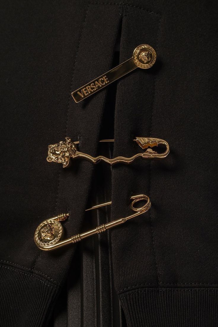 loseup shot of a black versace sweatshirt or hoodie. the garment features several gold safety pins adorned with the versace medusa logo, adding a unique and stylish design element.  one safety pin has the word "versace" engraved on it. the image highlights the texture of the fabric and the details of the metallic accents.