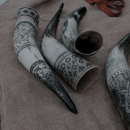 the image features a collection of four decorative drinking horns resting on a textured burlap fabric. the horns are likely made from animal horns and are adorned with intricate black and white engravings of various patterns and designs. a brown leather strap is also visible in the background. the overall setting suggests a historical or medieval context, showcasing the rustic and vintage aesthetic of the artifacts.
