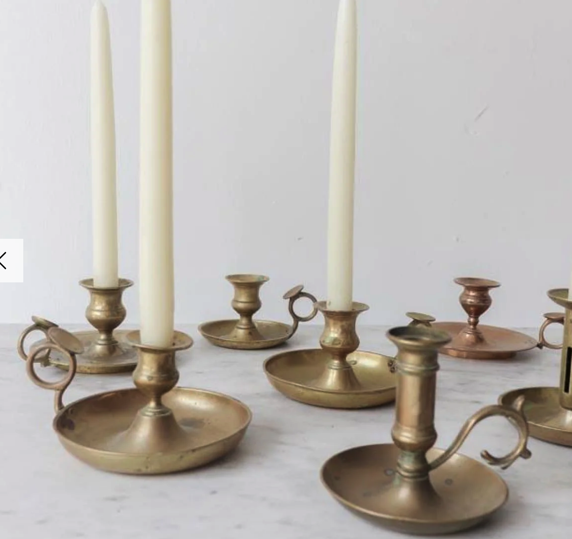 an image of multiple brass candle holders of different styles holding white candles. some are tall and slender while others are shorter and wider. there are a few of them with handles attached to them. the candles are in a variety of sizes, and all of them are lit. the image is taken from a high angle, and the background is a white surface.