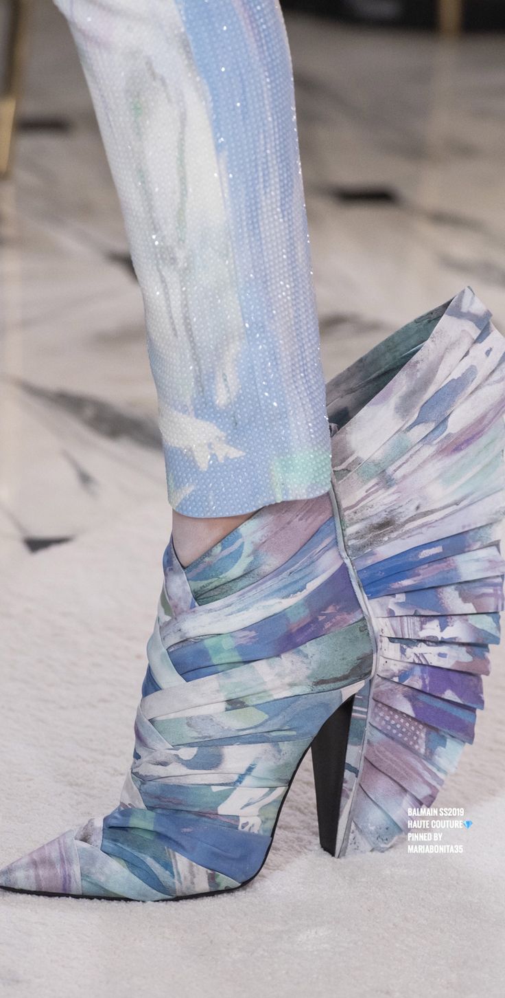closeup detail shot of a woman's right leg and foot showcasing a striking pair of balmain haute couture boots. the boots are highheeled stilettos with a pointed toe, featuring a unique, pleated abstract print in soft blues and pastels. the fabric appears to be a luxurious, possibly silk or satin blend, with a softly draped and textured design.  the boots are paired with light blue pants embellished with glitter or small beads, creating a cohesive and elegant look. the background is blurred, focusing attention on the shoes and the texture of the fabrics. the image suggests a runway or highfashion context, possibly from a fashion show.  a watermark indicates "balmain ss2023 haute couture," confirming the brand and collection.