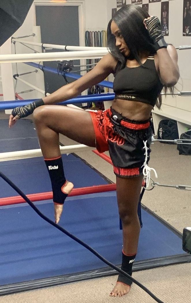 a female fighter wearing boxing gear and shorts stands in a boxing ring. she is in a fighting pose, demonstrating her athleticism and power.  she looks confident and determined.