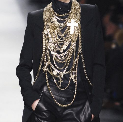 a model wearing a black leather jacket and pants with a gold cross necklace and a chain pendant. they have an elegant and sophisticated look.