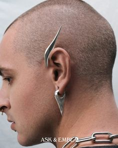 a closeup side profile shot of a person with a shaved head showcasing their ear piercings. they have two silver piercings: a  distinctive, pointed, winglike cuff on the upper ear cartilage and a geometric, triangular drop earring in their lobe. the individual has fair skin and a neutral expression. the soft lighting accentuates the contours of their face and highlights the metallic shine of the jewelry.