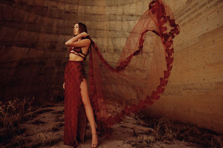 a woman in a long, flowing red dress poses inside a concrete silo. the dress has a thighhigh slit and a long, flowing train that billows out behind her. she is looking over her shoulder, and her hair is styled in loose waves. the lighting is soft and natural, and the overall effect is one of elegance and grace.