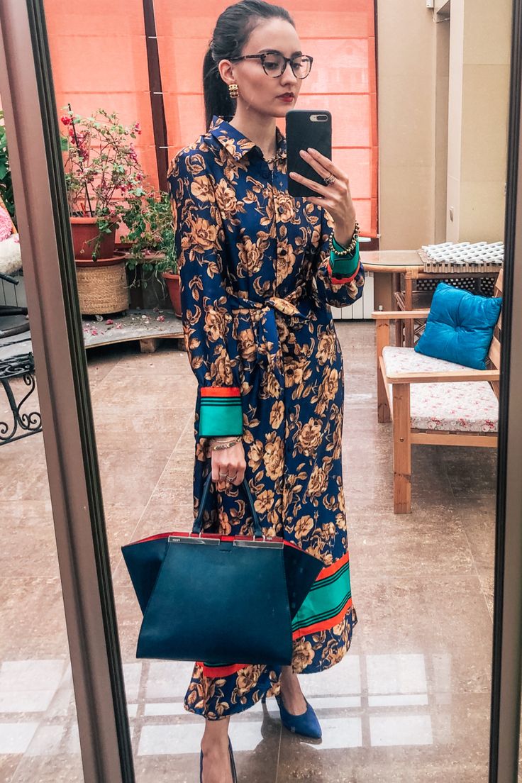 a woman in a stylish blue and gold floral maxi dress is standing in front of a mirror, taking a selfie.  the dress has colorful detailing on the cuffs and hem. she's carrying a navy blue tote bag and is wearing high heels and eyeglasses. the setting appears to be a home interior with plants and a patio visible through a glass door.