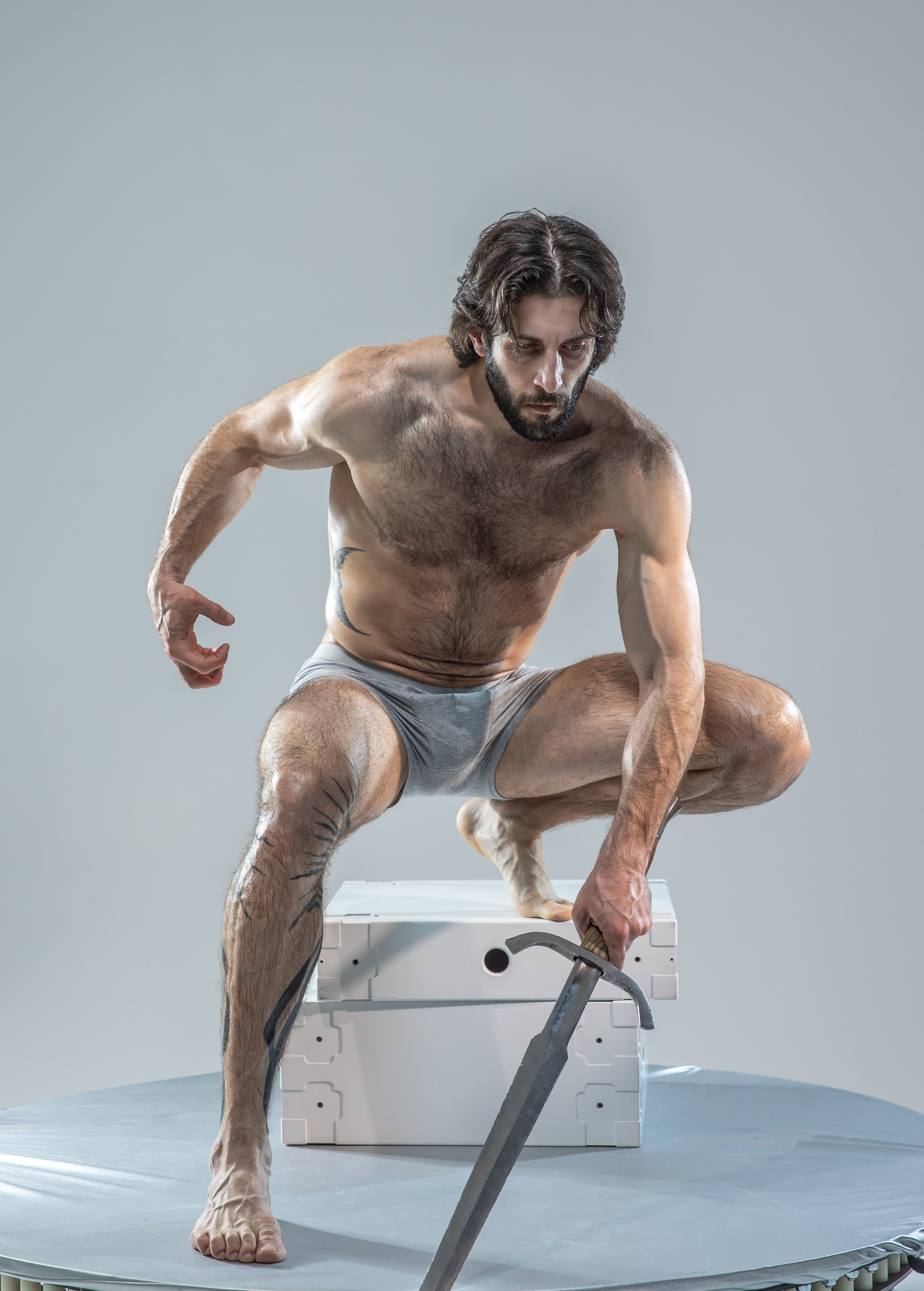 a muscular man with long dark hair and beard crouches on a white platform, holding a sword. he is wearing only gray underwear and has a tattoo on his left leg. the background is a light gray.