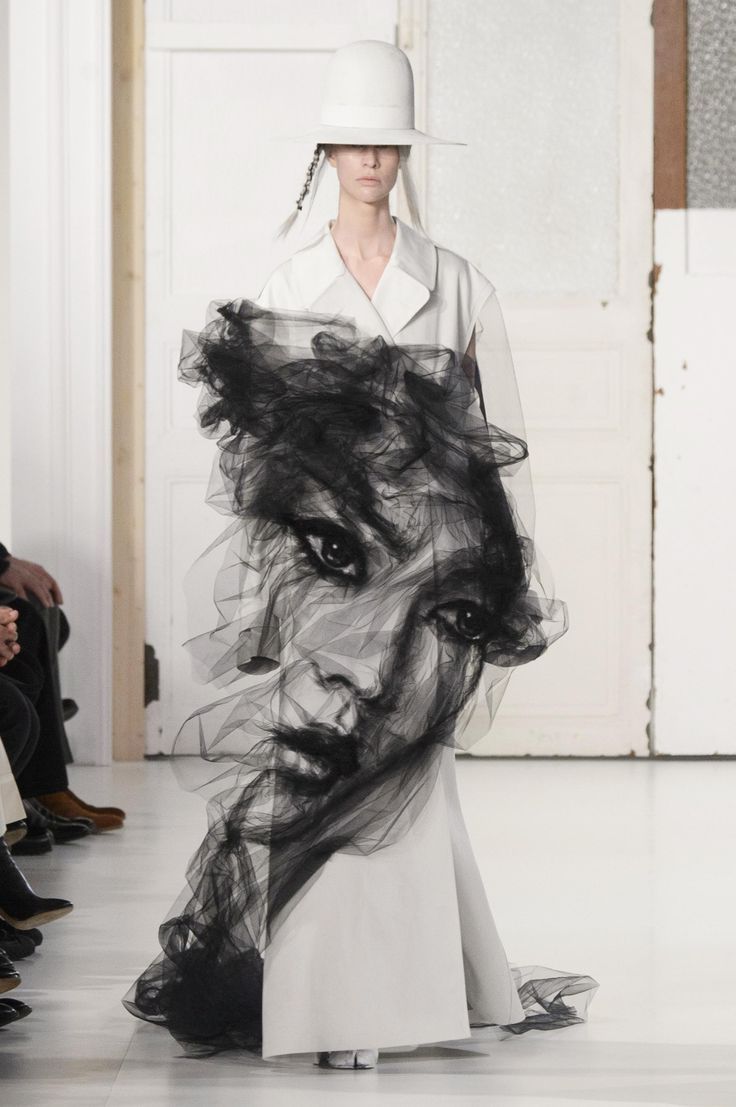 a model is walking on a runway wearing a white dress and a large white hat. the most striking feature of the outfit is a large, black tulle creation that forms a portrait of a face. the fashion show has white or lightcolored backgrounds. there are spectators to the side.
