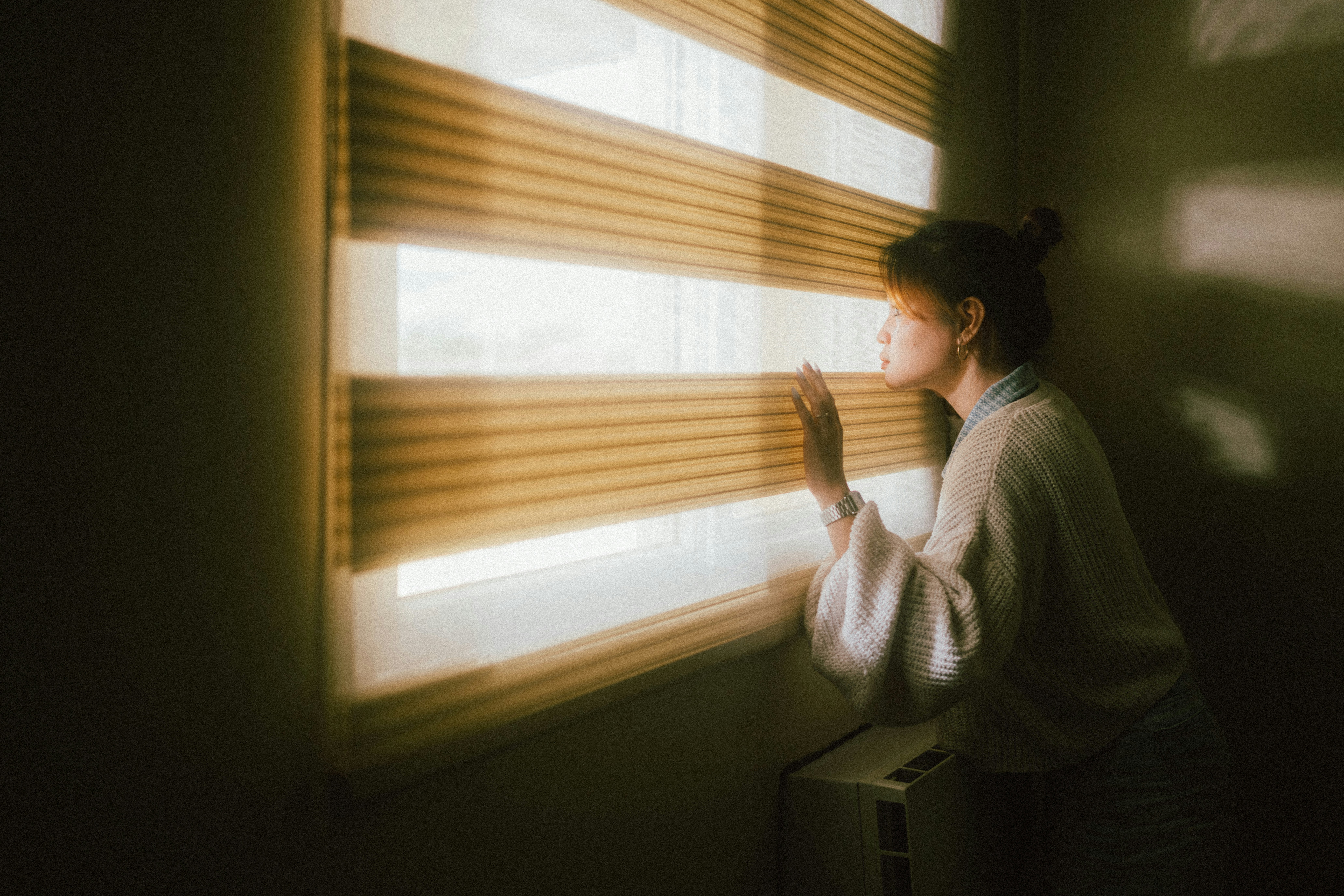 a woman stands by a window, her hand gently resting on the partially raised blinds. warm, natural light streams through the slats, illuminating her profile as she gazes out. the soft lighting and her contemplative pose create an atmosphere of quiet reflection or perhaps anticipation.