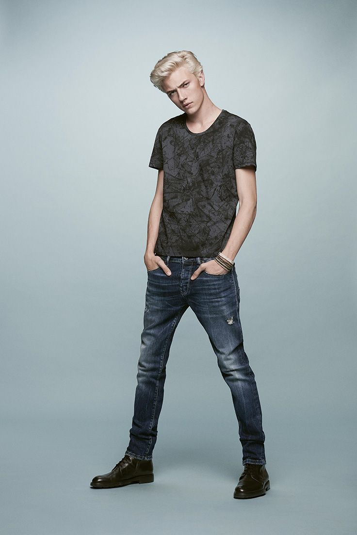 a full body studio shot of a young male model with blond hair wearing a black patterned tshirt, dark blue jeans, a bracelet, and dark brown dress shoes. he is standing with his hands in his pockets against a light blue background.
