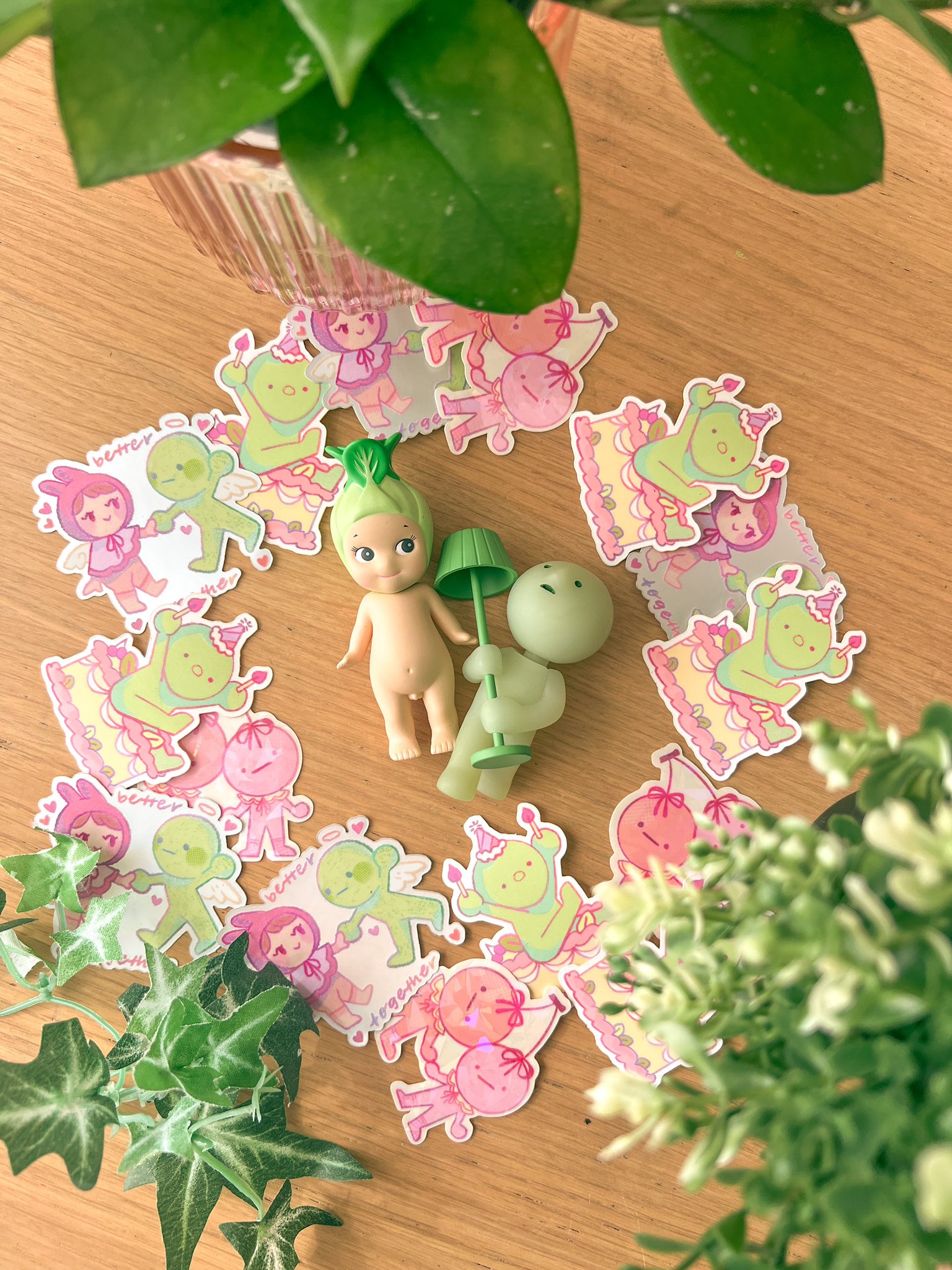 he image is a flatlay photograph showcasing a collection of cute, pastelcolored stickers featuring chibistyle vegetable characters. the stickers are arranged in a circle on a light brown wooden surface. two small figurines, one resembling a baby bok choy and the other a small amorphous creature, are centrally placed among the stickers.  lush green potted plants are prominently featured in the foreground and background of the image, adding to the overall bright and cheerful aesthetic. the overall style is kawaii, whimsical and playful.