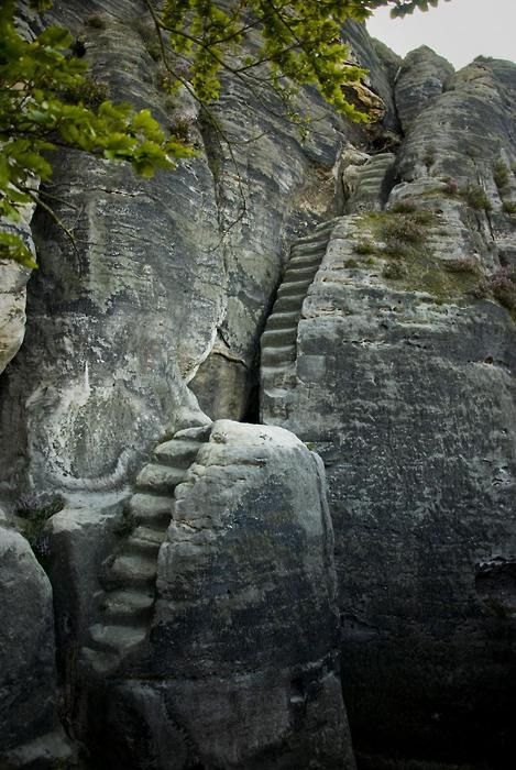 a view of stone steps carved into the side of a large rock formation. the steps lead upwards into the rock, suggesting a path for climbing or exploration. the rock formations are made of layered sandstone, and some greenery is visible near the top of the rock. the overall image suggests an outdoor adventure or hiking trail through a mountainous landscape.