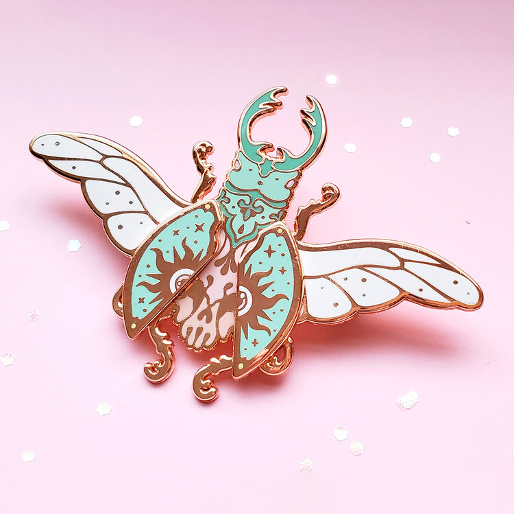 the image shows a detailed enamel pin in the shape of a beetle. it features elaborate designs with white wings lined in gold, a blue body with decorative patterns, and other gold details, on a light pink background with glitter specks.