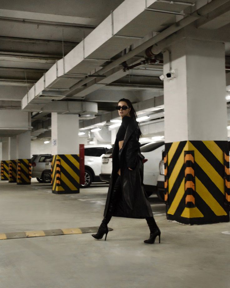 a woman wearing a black coat, boots, and sunglasses poses in a parking garage. she is standing confidently with her legs slightly apart and her arms at her sides. the walls of the garage are lined with yellow and black striped caution tape.
