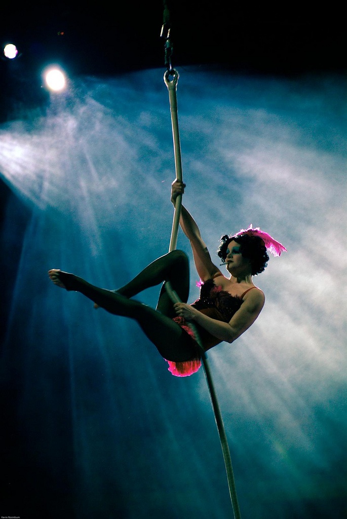 a woman in a black costume and a pink feathered headdress hangs from a trapeze rope. she is positioned on a stage bathed in blue light and surrounded by smoke. her legs are straight, creating a silhouette against the blue, and she is looking down. the image is taken from a low angle.