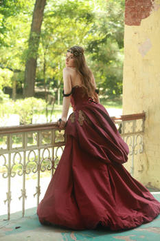 a woman in a long, burgundy gown stands on a porch overlooking a garden.  the dress is a full, flowing style with a somewhat historical or vintage look. the woman has a crown on her head and her hair is styled to cascade down her back. the setting is outdoors in a garden with trees and a partially ruined building in the background. she is leaning on a white wroughtiron railing. the overall feel is romantic and elegant.