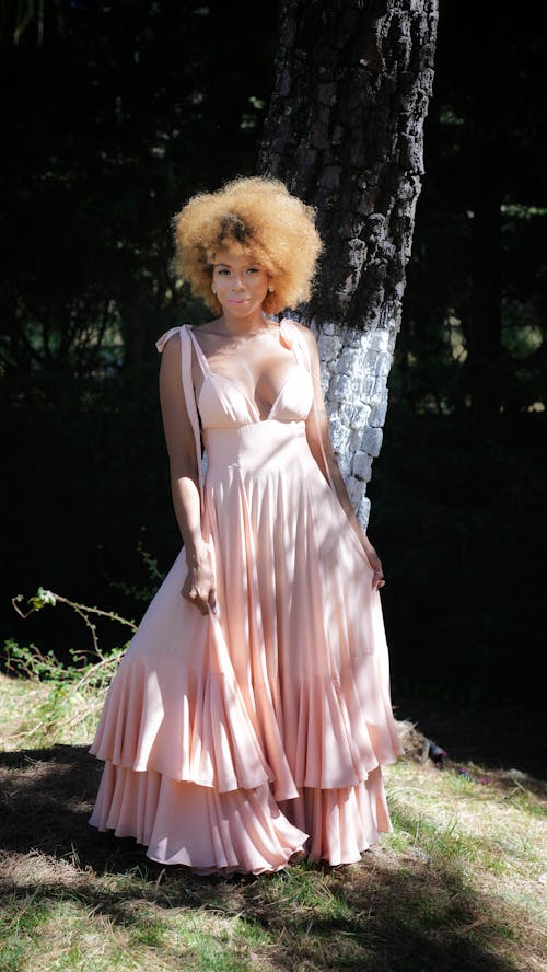 a beautiful woman with an afro hairstyle stands outdoors in a pink maxi dress.  the dress is layered with ruffles and has thin straps. she's smiling gently and appears happy. the setting is a wooded area with a large tree trunk behind her.  the sunlight casts shadows on the dress and the surrounding grass.