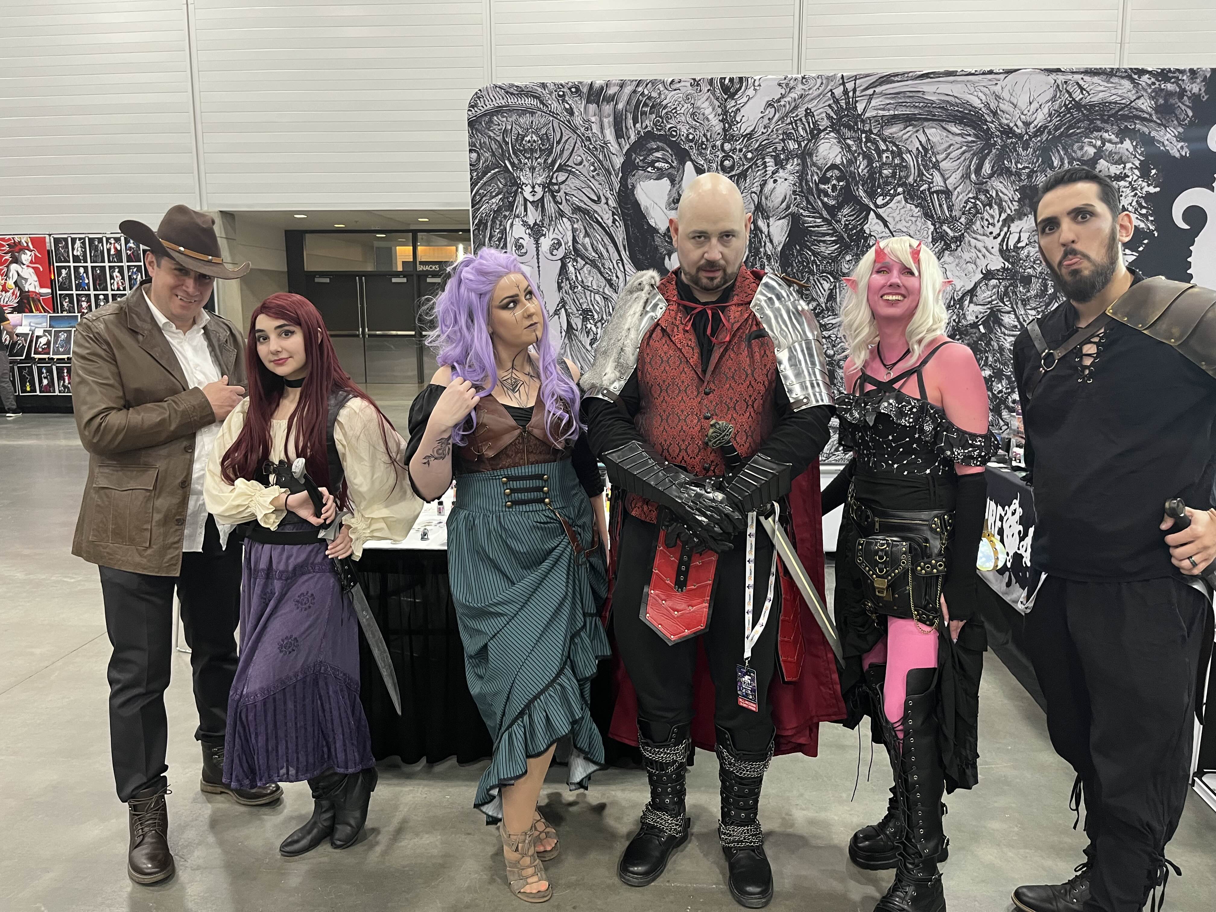 a group of six cosplayers, dressed in elaborate fantasy costumes, pose for a photograph at a convention. the costumes include steampunk and medieval elements, with a dark fantasy aesthetic.  one person is dressed as a gunslinger, another in a pirateinspired outfit, others in more elaborate, ornate attire. the background shows a large banner with dark fantasy artwork. the group appears to be having fun and enjoying the event.