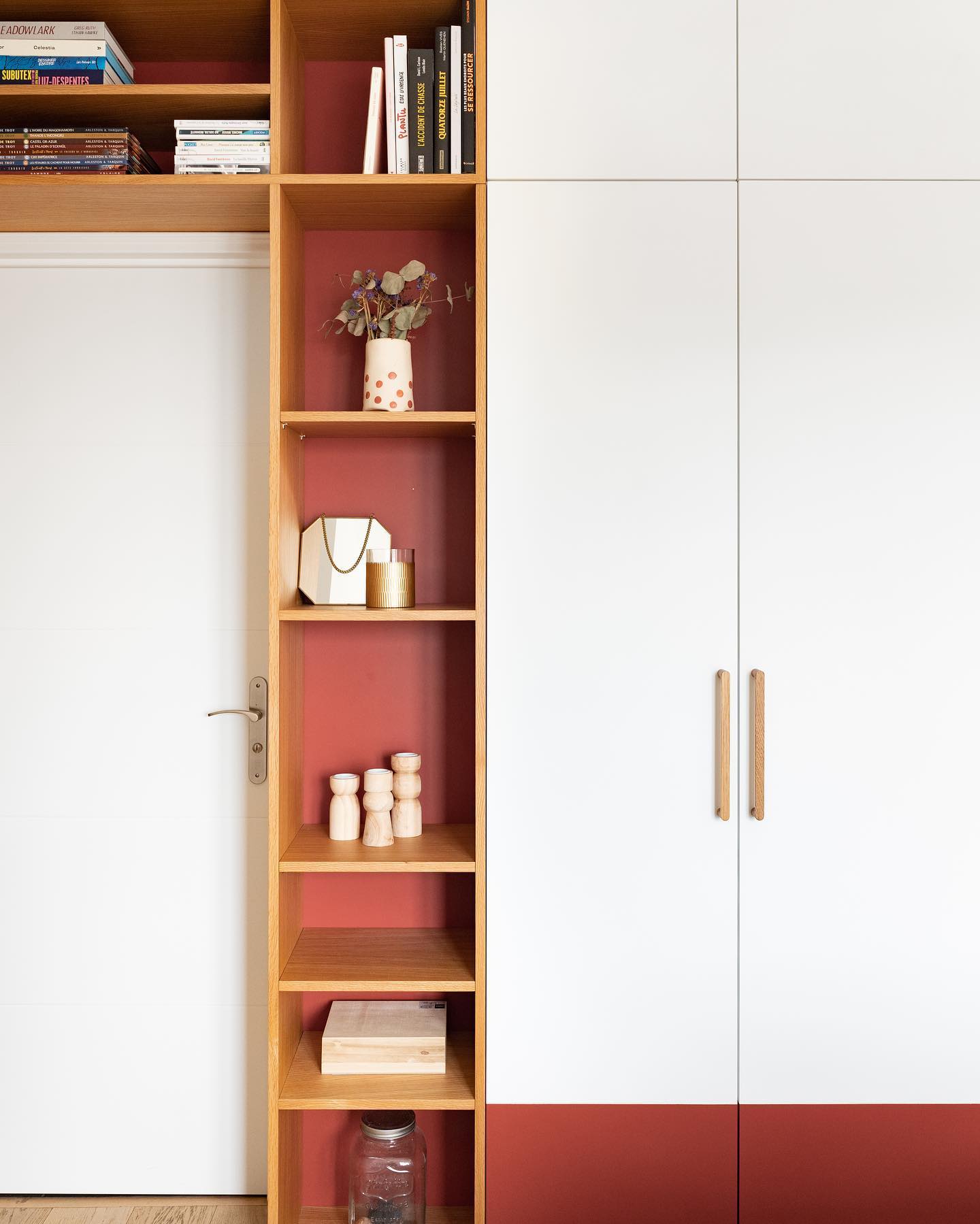 image shows a white door adjacent to a builtin bookcase and wardrobe unit. the bookcase has a light brown wood frame with red painted backs to the shelves. it holds various items including books, a vase with dried flowers, a handbag, a candle, wooden figurines, a box, and a glass jar. the wardrobe has white doors with light brown wooden handles and a red accent at the bottom.