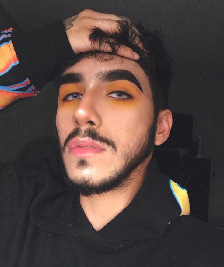 a portrait of a man with a beard and blue eyes, wearing makeup, with his hand in his hair and wearing a hoodie.