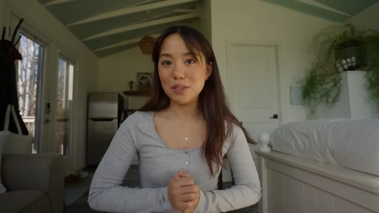 a young woman is standing inside a tiny home. she is looking at the camera and talking. she is wearing a gray shirt and has long black hair. the interior of the tiny home is bright and airy with white walls and light wood accents. there is a bed in the background and a living room area in the foreground.