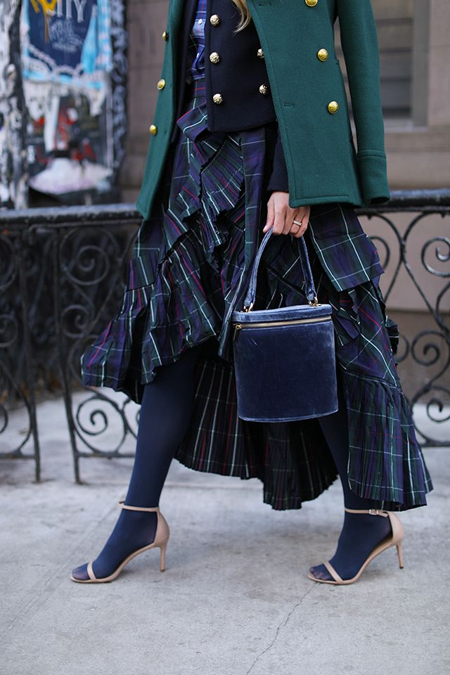 a woman wearing a stylish autumnal outfit.  she's wearing a dark green peacoat over a plaid highlow skirt, paired with navy tights and beige ankle strap heels. she carries a dark blue velvet bucket bag and accessorizes with gold jewelry. the photo is taken outdoors on a city street.