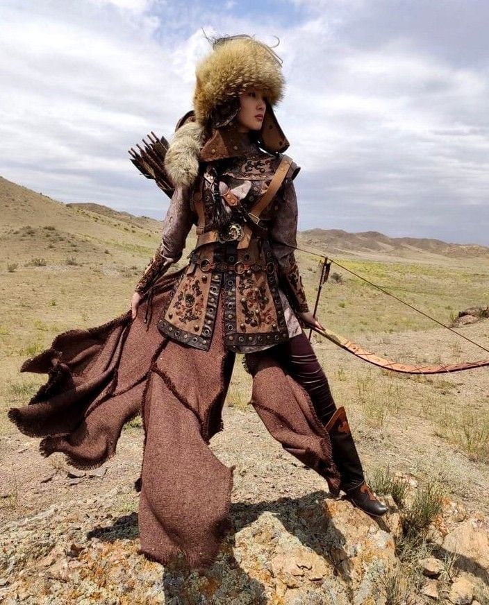 a woman dressed in traditional central asian attire poses with a bow and arrow in a desert landscape. she is wearing a large fur hat, an intricately decorated leather vest and skirt, and tall boots. she is holding a bow and arrow and there is a quiver of arrows on her back. the landscape behind her is a dry, hilly terrain with a cloudy sky.