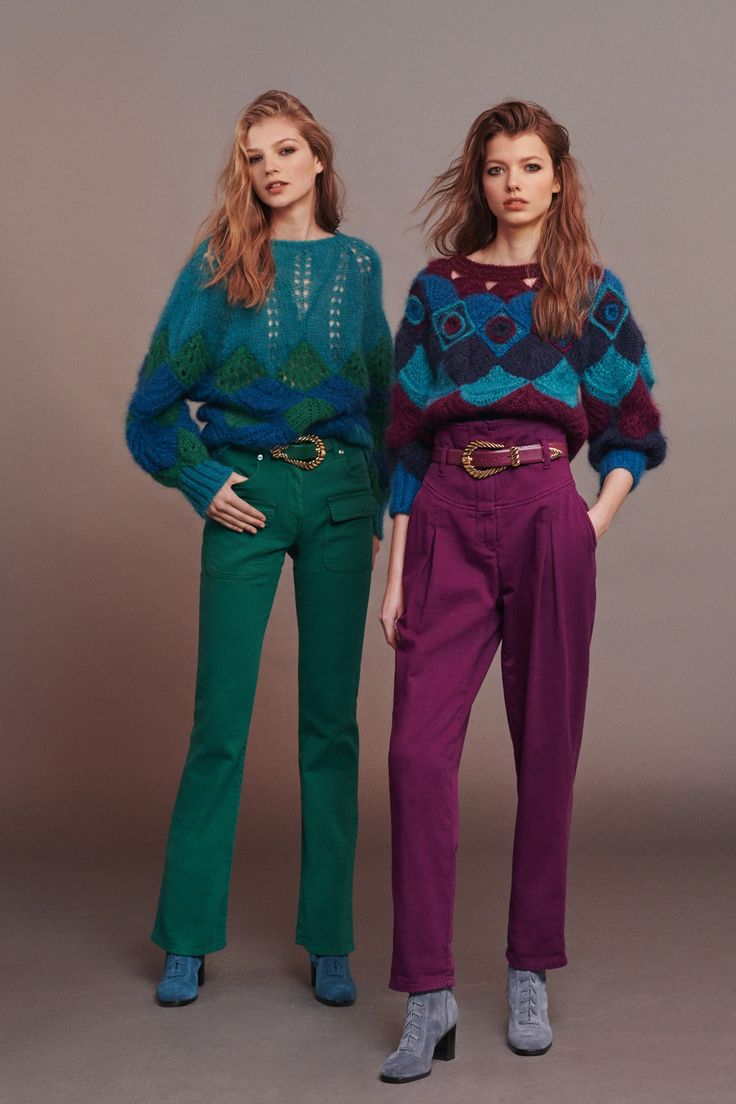 two female models are standing against a neutral gray background. they are both wearing colorful patterned sweaters and highwaisted pants of different colors. one model is wearing emerald green pants, while the other is wearing burgundy pants. they're also wearing matching light blue ankle boots. the sweaters have a retro, slightly bohemian style. the overall image is a fashion photoshoot showcasing the latest clothing collection.