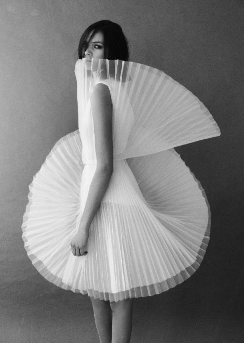 a black and white image of a woman in a white, pleated dress with a ruffled collar. she is standing with her arms down, looking towards the side of the image. the background is a plain wall, giving the woman and her dress more emphasis.