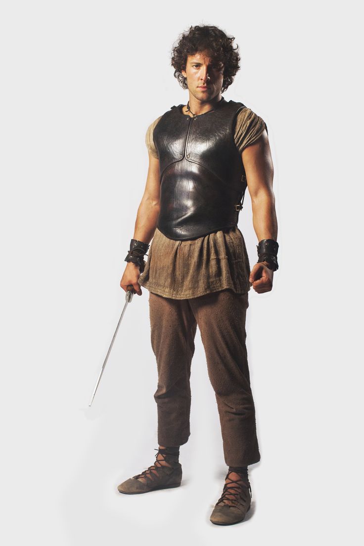 a man stands in a costume. he wears a leather vest, arm cuffs, tunic, pants and shoes. he also holds a sword.