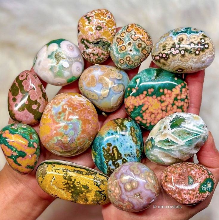 a hand is holding a variety of ocean jasper stones, each one with unique color patterns and textures.  they are all tumbled and polished smooth, giving them a beautiful shine. ocean jasper is a gemstone known for its spiritual and healing properties. it is believed to promote peace, harmony, and grounding.  it is also said to be a powerful stone for clearing negative energy and opening the heart chakra.  this image captures the beauty and magic of ocean jasper.