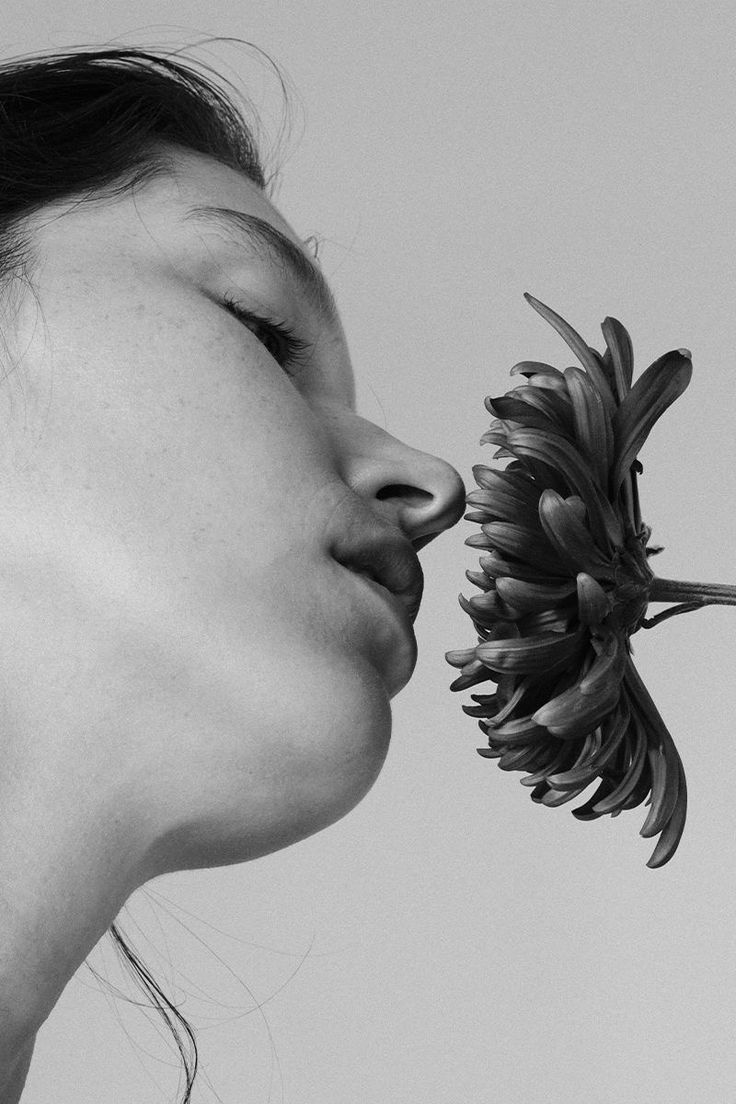 a black and white closeup portrait of a woman's face in profile, her eyes closed as she gently inhales the scent of a darkcolored gerbera daisy held near her nose. the image is softly lit, creating a serene and elegant mood.  the focus is sharp on the woman's face and the flower, with a softly blurred background. the overall aesthetic is minimalist and clean.