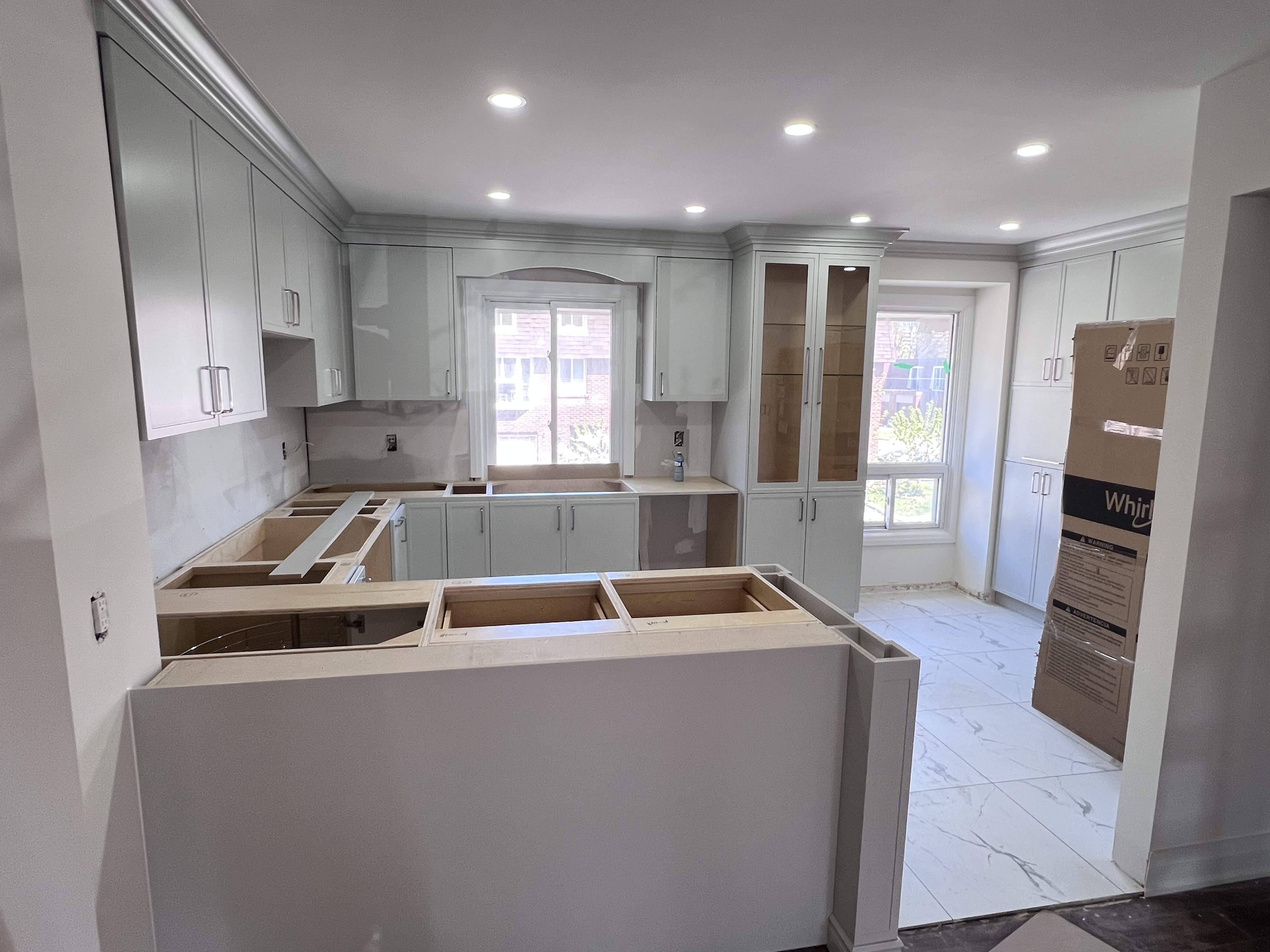 the image shows a kitchen that is under construction. the upper cabinets are installed, but the countertops are missing. the kitchen island is built, but unfinished. there is a box with a whirlpool logo in the background. the floor is tiled.