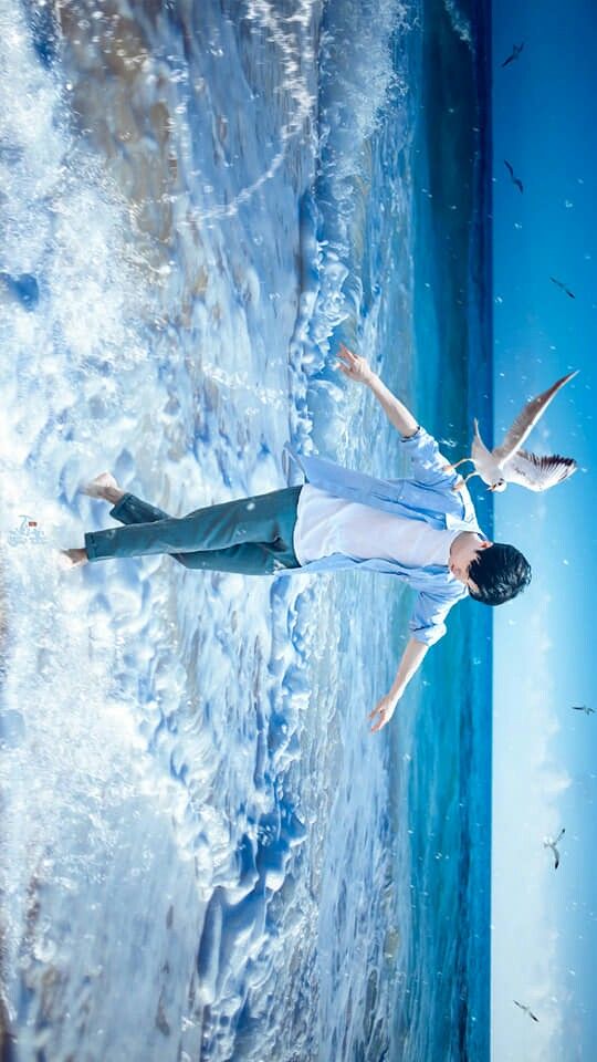 a young man with short black hair is captured midstride in shallow ocean water. he wears a light blue, loosefitting shirt and gray pants, his arms outstretched as if embracing the vastness of the sea. white seagulls soar around him against a vibrant blue sky, while frothy waves crash at his feet. the scene evokes a sense of joy, freedom, and connection with nature.