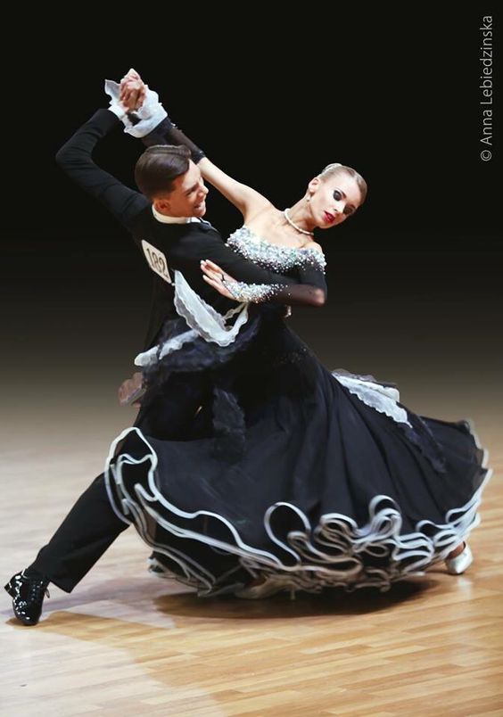 a man and a woman are ballroom dancing on a wooden dance floor. the woman is wearing a black and white ball gown and the man is wearing a black suit. the woman's dress is flowing and she is holding the man's hand. the man is looking at the woman and the woman is looking away. the background is black.