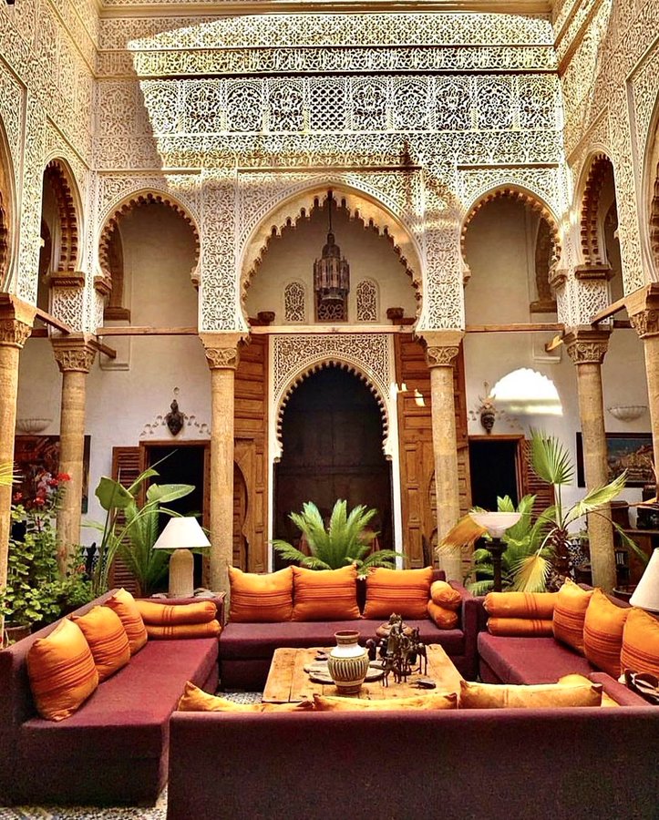 the image shows an intricately designed moroccan courtyard interior. the walls feature ornate carved arches and detailed architectural elements. columns support the arches, framing the space. in the center of the courtyard, there is a comfortable seating area with a sofa adorned with colorful cushions and pillows. a wooden table sits in front of the sofa, holding various decorative items. plants are placed throughout the courtyard, adding a touch of nature. a doorway leads into another area, and a lamp provides soft lighting. the overall style is reminiscent of traditional moroccan design, with a focus on detail, craftsmanship, and a warm, inviting atmosphere.