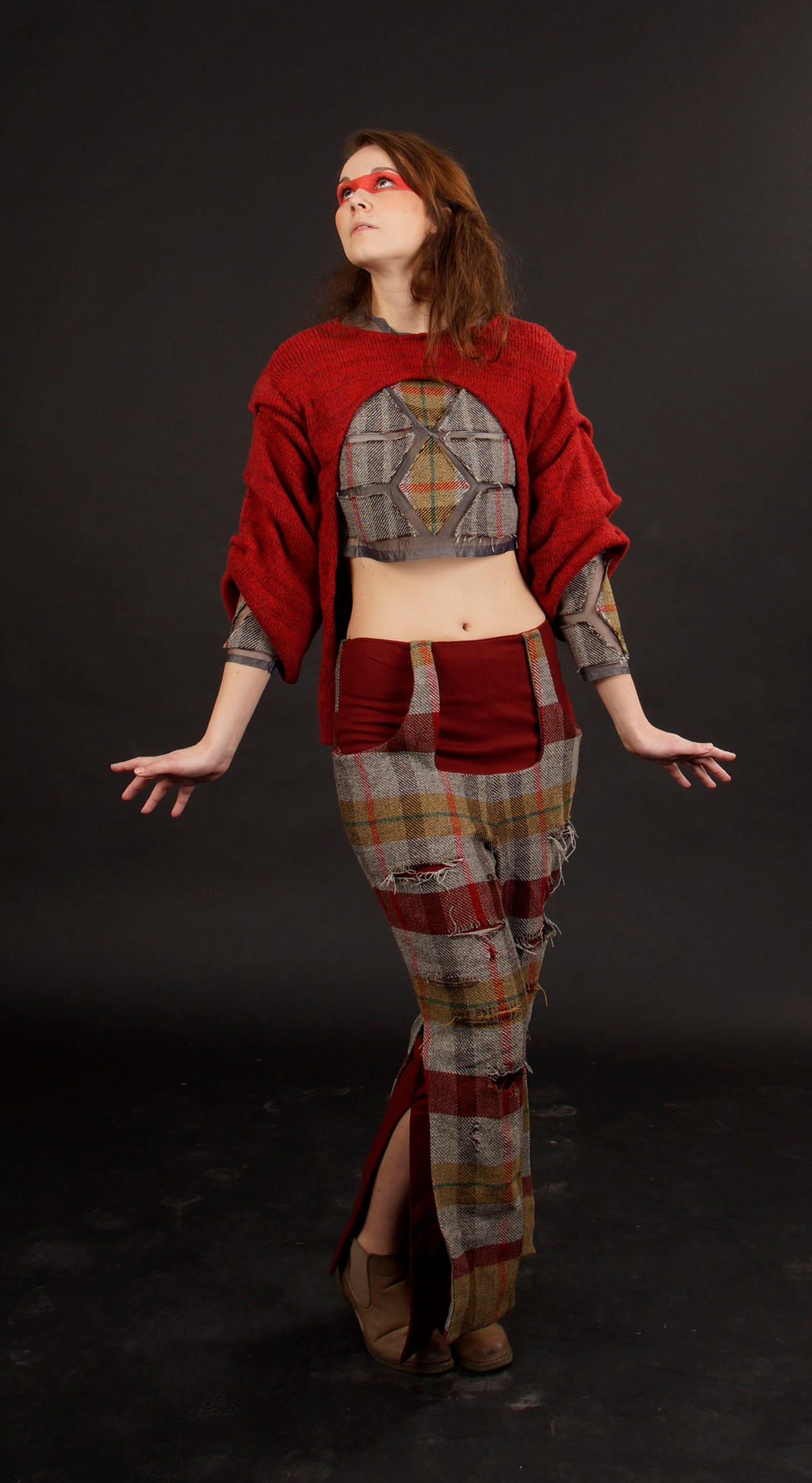 a fulllength studio shot of a female model showcasing a unique outfit. the model has red markings under her eyes and is looking upward. she is wearing a loosefitting red sweater with a patched plaid bodice and highwaisted, patchwork pants in shades of red, beige, and brown.  the pants have distressed areas showing the underlying fabric.  she is wearing brown ankle boots. the background is a simple, dark gray. the overall style is artistic and unconventional.