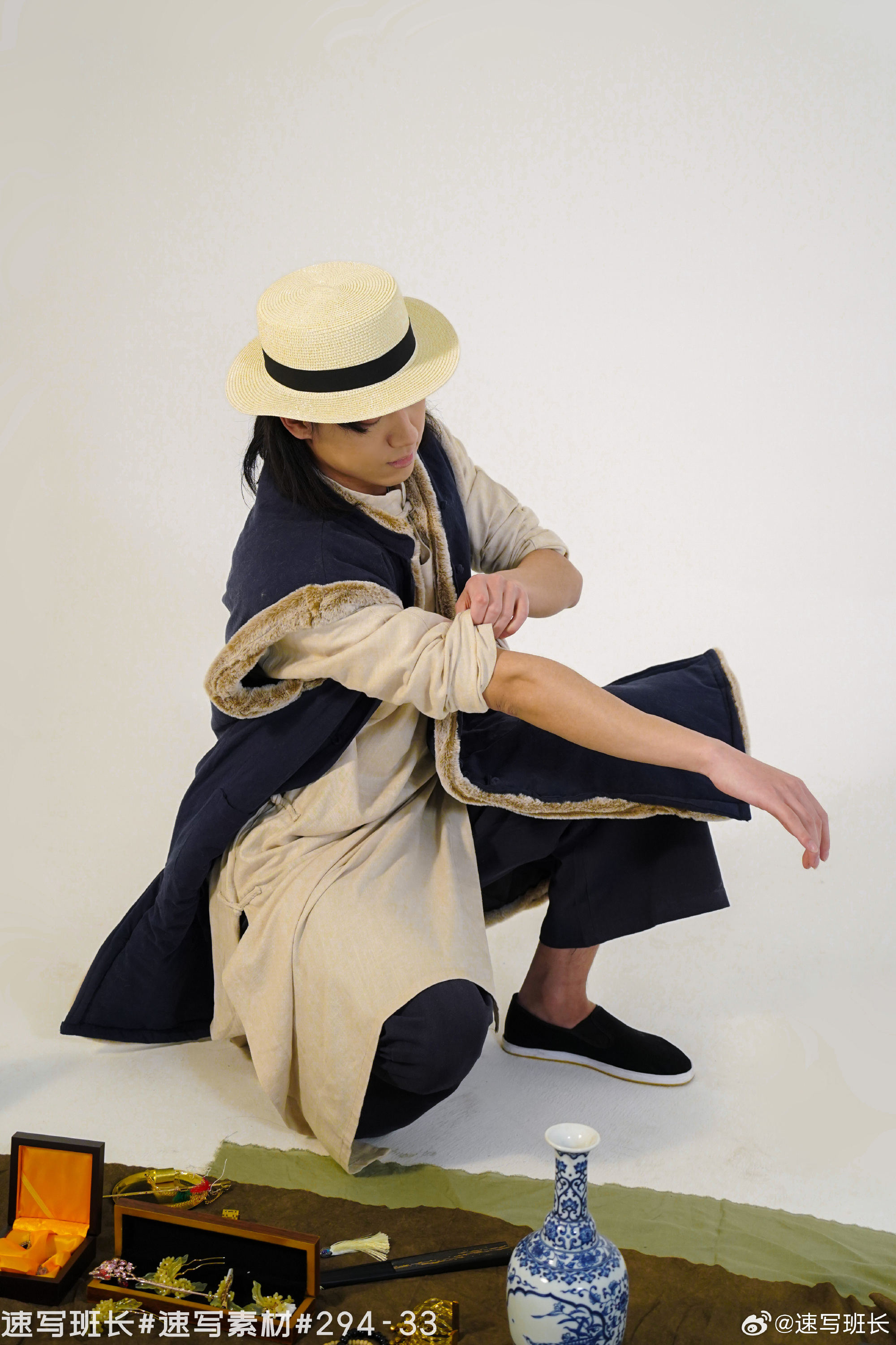a young man with long black hair, wearing a straw hat, is kneeling and adjusting the sleeve of his traditional chinese outfit. the outfit consists of a long, beige robe with a blue, furtrimmed vest. he is wearing dark blue pants and black cloth shoes. in the foreground are a blue and white porcelain vase, a wooden box with gold accents, and other decorative items. the background is a plain white studio backdrop.