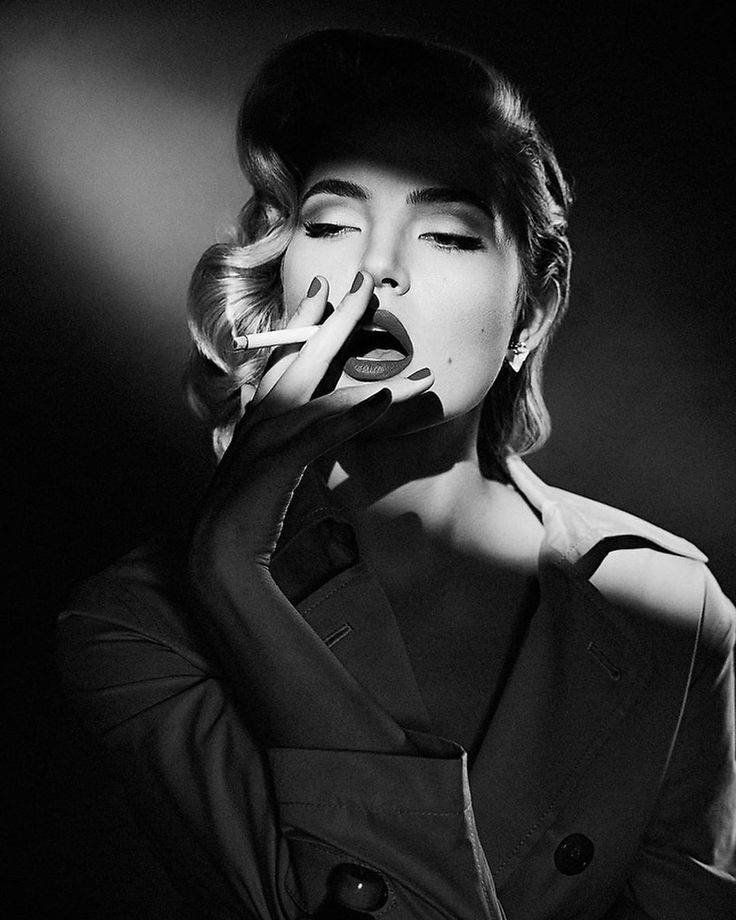 a monochrome portrait of a glamorous woman with wavy hair and a coat, smoking a cigarette. she has red lipstick and a sensual expression.