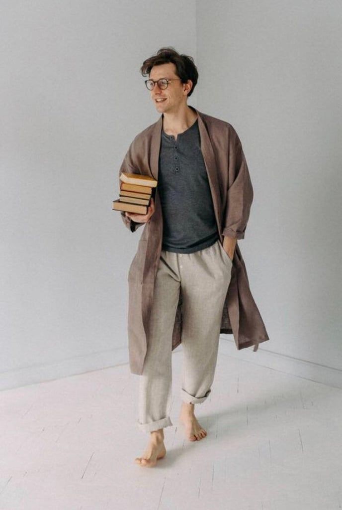 a full shot of a smiling, handsome man wearing a brown linen robe and beige pants, casually holding a stack of books.  he is barefoot and walking across a lightcolored floor in a minimalist, bright room. the overall style is relaxed and comfortable.