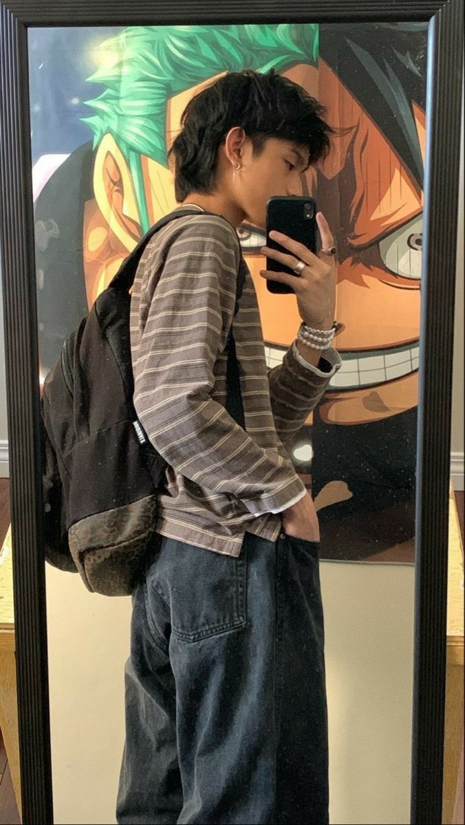 a man is taking a mirror selfie. he is wearing a striped shirt, jeans, earrings, bracelet, and ring. he has a backpack on. behind him is an anime poster of zoro from one piece.