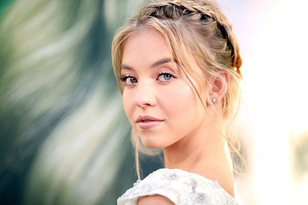 the image is a closeup portrait of a young blonde woman with her hair styled in a partial braid. she is wearing light makeup and a white, embellished dress, suggesting a formal occasion like a wedding or red carpet event.  her expression is soft and she is looking over her shoulder towards the viewer. the background is blurred, drawing attention to the woman. the overall mood is elegant and beautiful.