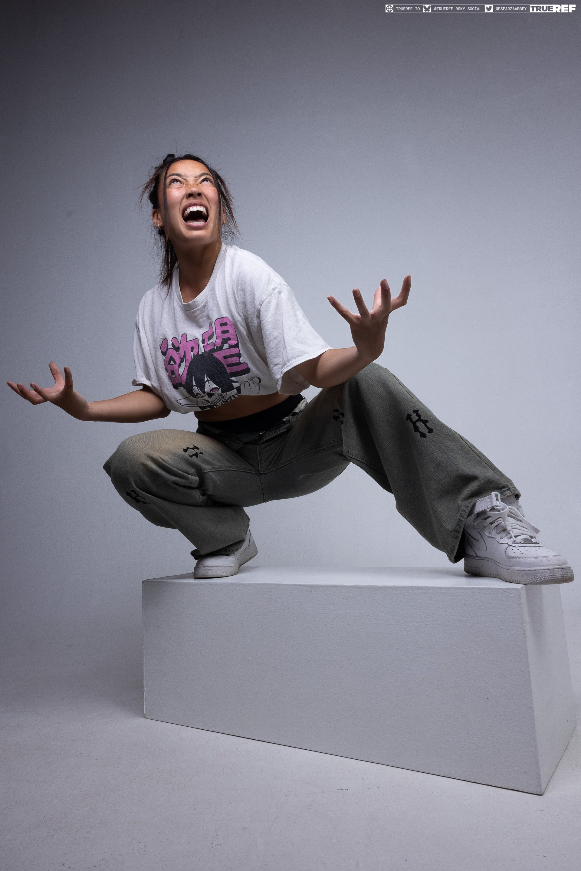 a woman in a white tshirt with an anime graphic, khaki pants with black designs, and white sneakers is crouching on a white platform. she has her hands raised and mouth open as if yelling. the background is a plain studio backdrop.