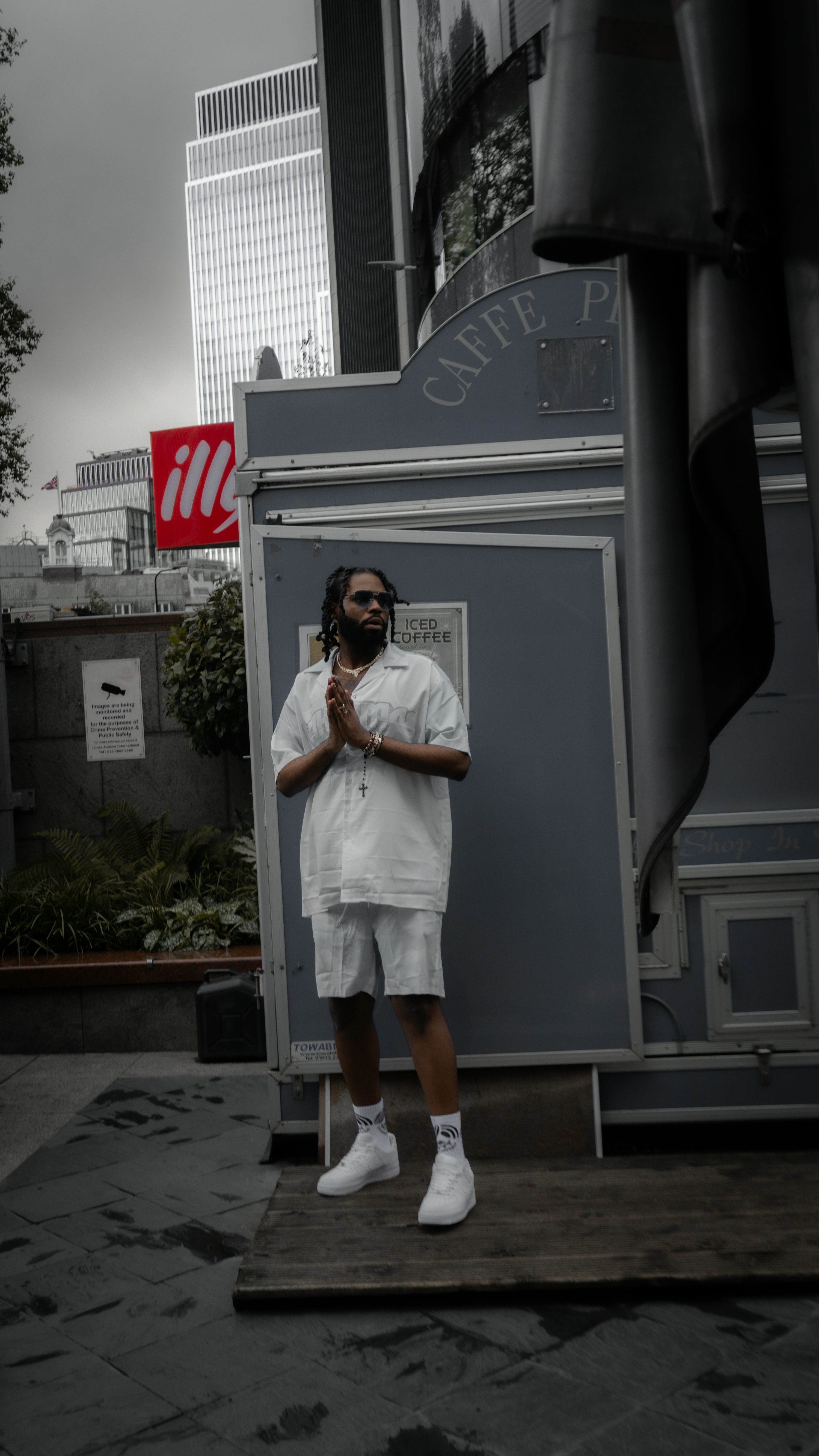 a fullbody shot of a young man standing in front of a gray coffee shop kiosk in an urban setting. he's dressed in a white shortsleeved shirt and matching white shorts, paired with white air force 1 sneakers. his hands are clasped together in a praying position. the background features modern city buildings, including tall skyscrapers and a prominent illy coffee sign. the overall atmosphere is moody and dark, contrasting the bright white of the model's outfit. the image has a strong fashion editorial feel.