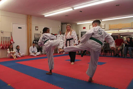 a group of people wearing taekwondo uniforms practice kicks and other martial arts techniques in a gym with a red and blue mat.  a young woman in a black belt uniform is instructing two students.  several other people are watching the class.