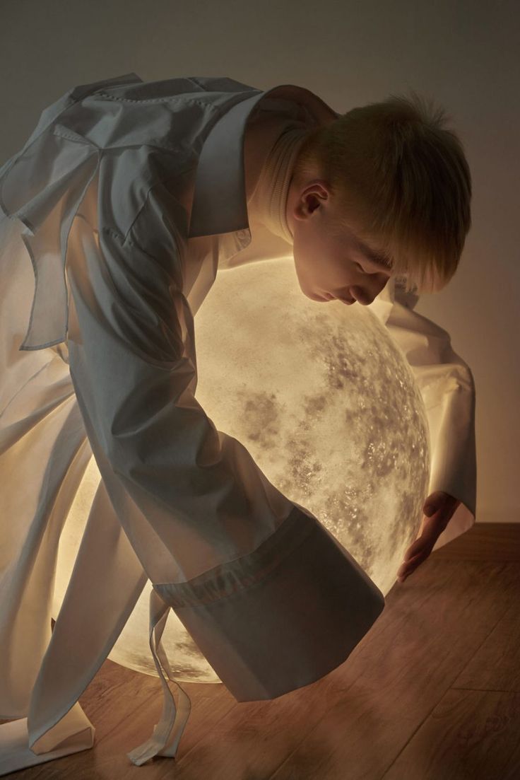 a young, blonde male model, dressed in an oversized white shirt, is captured in a closeup shot, interacting with a large, illuminated moon prop that dominates the lower half of the frame. he leans over the moon, his face close to its surface, his hands gently resting on its curve. the warm, artificial light emanating from the moon casts soft shadows and highlights on the model's face and clothing. the background is a neutral, undefined brown, keeping the focus on the model and the ethereal moon.
