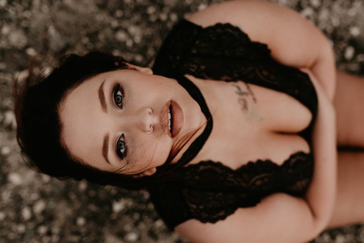 the image is a closeup, overhead shot of a plussize woman lying on the ground outdoors. she's wearing a black lace bodysuit and has a script tattoo on her chest. her makeup is noticeable, and she's looking directly up at the camera with a sensual expression.  the overall mood is intimate and confident, showcasing body positivity. the background is blurred, focusing attention on the woman.