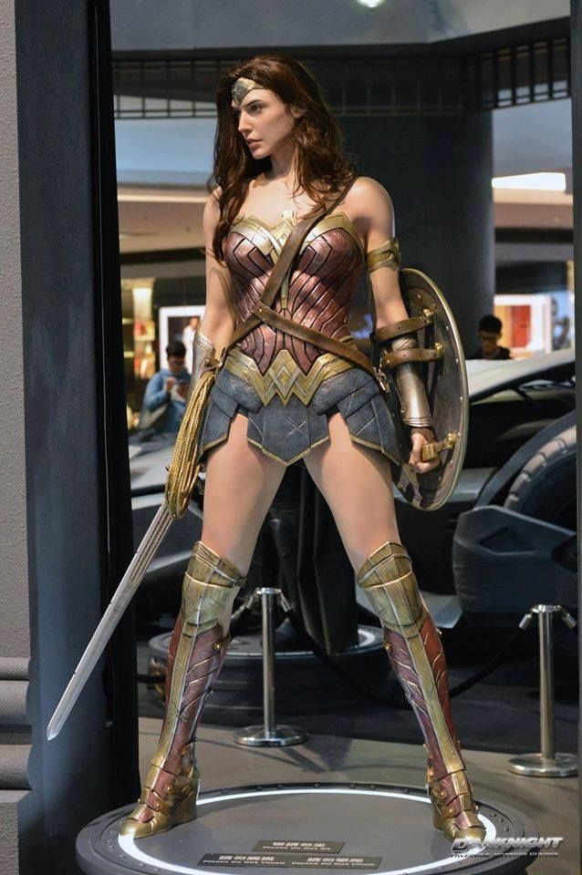 a wonder woman action figure stands on a display platform with her sword and shield. she is wearing her signature costume, including the red, gold, and blue armor and her iconic tiara. the figure is positioned in a dramatic pose, as if she is ready for battle.  she is standing in front of a large, black car,  which helps to emphasize her size and power. the background of the image is a blurry mix of lights and shadows, giving the photo a sense of depth and intrigue.